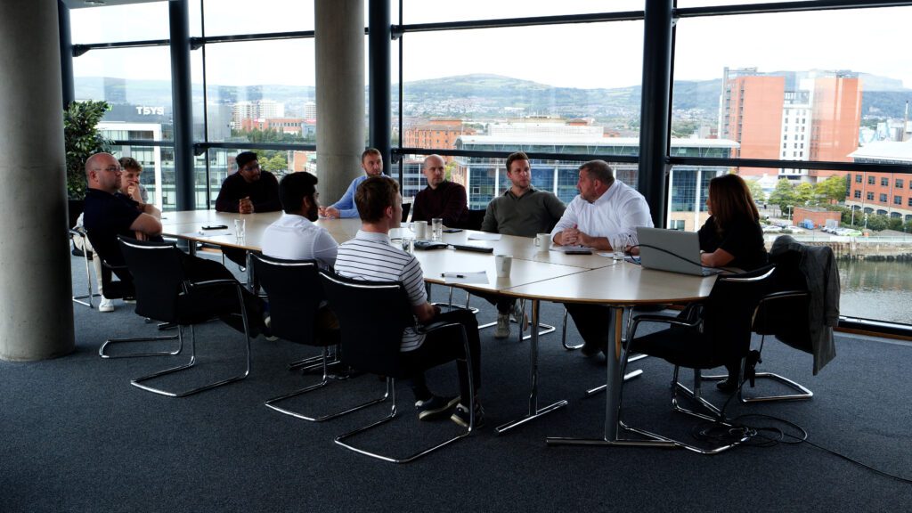 A group of people sit around a large conference table in a modern office with large windows overlooking a cityscape and river, preparing for a Business Eye interview.