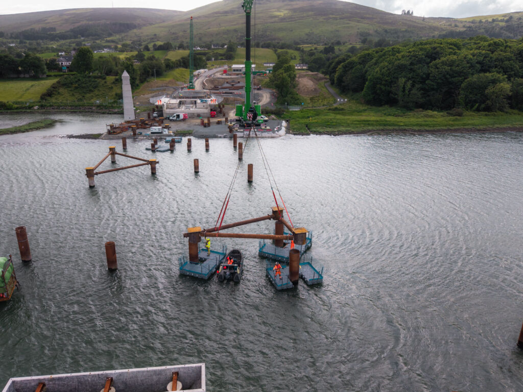 Crane lifting a steel bracing frame into position over temporary piles in the River Newry as engineers work from floating pontoons